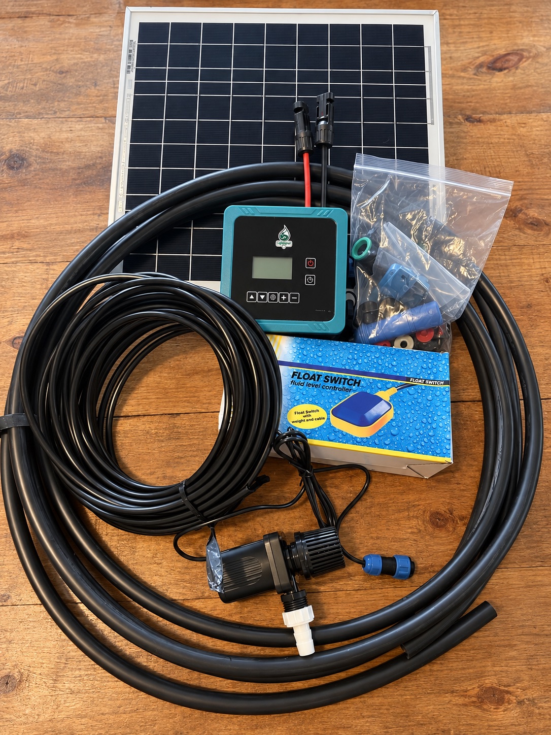 Greenway Drip solar irrigation components arranged on a wood table, including the solar panel, programmable controller, pump, tubing, float switch, and fittings